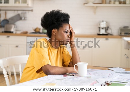 Bored young african american girl tired of remote study or freelance work sit at home workplace in kitchen look aside. Apathetic black female architect designer frustrated suffer from pain or stress
