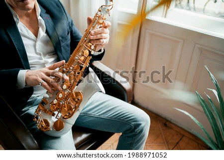 An adult gray-haired male musician artist sits at home and holds a saxophone in his hands.