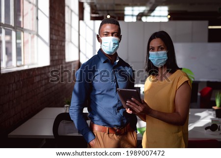 Portrait of diverse male and female colleague wearing face masks standing in office using tablet. working in business at a modern office during coronavirus covid 19 pandemic.