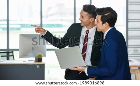 Side view of Asian male entrepreneurs standing with laptop in workplace and discussing project while pointing away