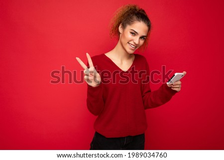 Photo of happy smiling woman with gathered curly hair wearing dark red sweater isolated over red background holding smartphone having fun and showing peace gesture looking at camera