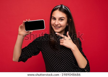 Closeup photo of happy Pretty brunette girl standing isolated over red wall wearing casual stylish black clothes showing mobile phone with empty screen for cutout looking at camera and pointing finger