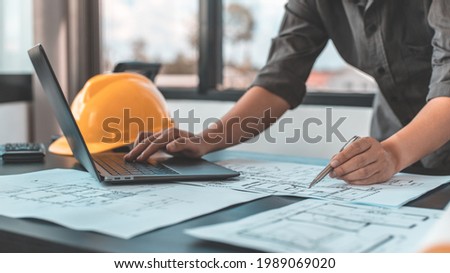 Young man with a laptop plotting a system of building structures in blueprints, Architects or engineers are designing buildings using computers to calculate the physical structure to be correct. Royalty-Free Stock Photo #1989069020