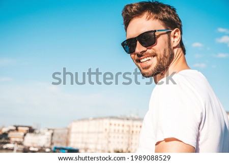 Closeup portrait of handsome smiling  hipster lambersexual model.Stylish man dressed in white T-shirt. Fashion male posing behind blue sky on the street background in sunglasses Royalty-Free Stock Photo #1989088529