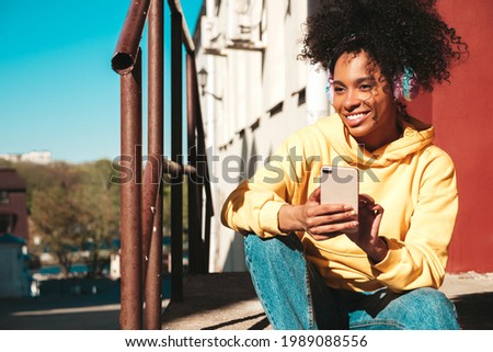 Beautiful black woman with afro curls hairstyle.Smiling model in yellow hoodie.Sexy carefree female enjoying listening music in wireless headphones.Posing on street background at sunset.Holds phone Royalty-Free Stock Photo #1989088556