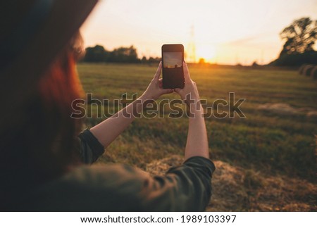 Stylish woman taking photo of sunset on phone in summer field. Young female holding smartphone and capturing evening warm sunshine in countryside. Atmospheric beautiful moment