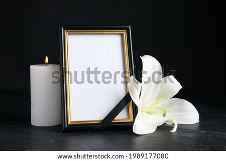 Funeral photo frame with ribbon, white lily and candle on black table against dark background. Space for design
