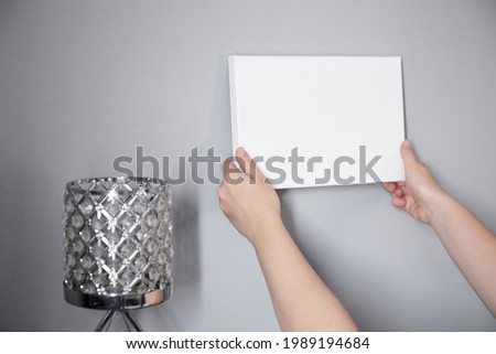 White canvas in female hands with gray wall background. Woman hanging blank picture mockup on wall. Wall decor. Canvas stretched on frame