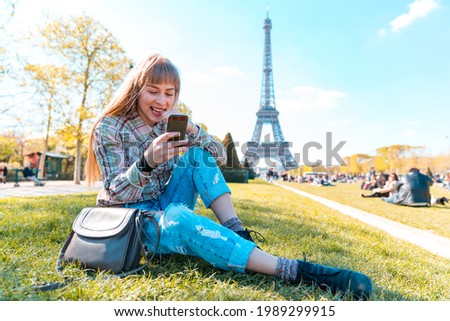Happy girl using phone in Paris with Eiffel Tower on background - Portrait of happy blonde young woman sitting on the grass and looking at her smartphone - Travel and lifestyle concepts