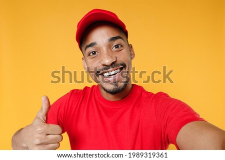 Close up fun delivery employee african man 20s in red cap blank print tshirt uniform workwear work courier dealer service concept doing selfie shot on mobile phone isolated on yellow background studio