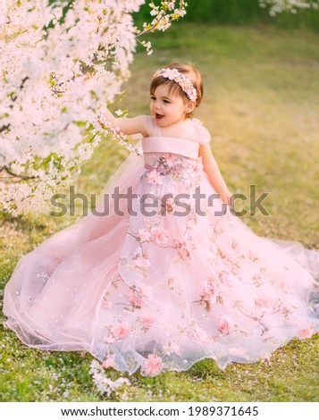 Young happy little girl fairy princess. Happy child face is smiling. Luxurious fluffy long dress for children, pink outfit. White flowers background trees spring nature. Fashion model, one year old