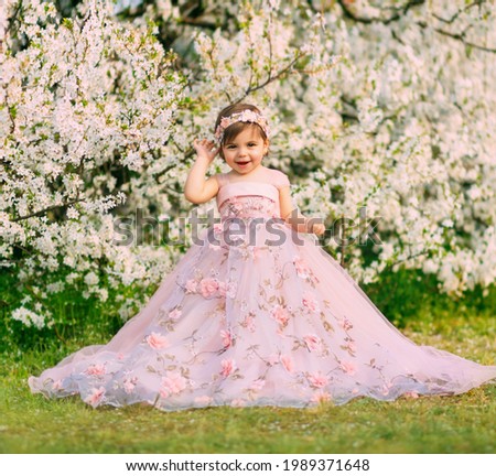 Young happy little girl fairy princess. Happy child face is smiling. Luxurious fluffy long dress for children, pink outfit. White flowers background trees spring nature. Fashion model, one year old
