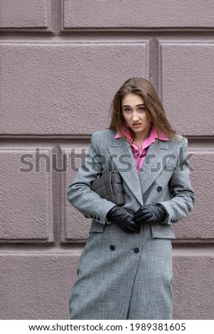 A vertical photo of a young girl with a cocky look and a clear emotion of indignation on her face, wearing a gray coat and a pink shirt with black leather gloves outside against a wall.