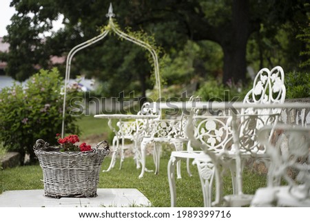 empty green wedding garden with red flowers in wicker basket and white Victorian metal decorative table and chairs