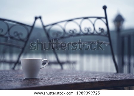 closeup detail of white small espresso coffee cup on wooden table with rain drops on rainy outdoor restaurant terrace 