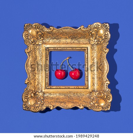 Bright red cherry and retro golden painting frame on bold blue background. Spring or summer aesthetic fruit creative idea. Minimal flat lay.