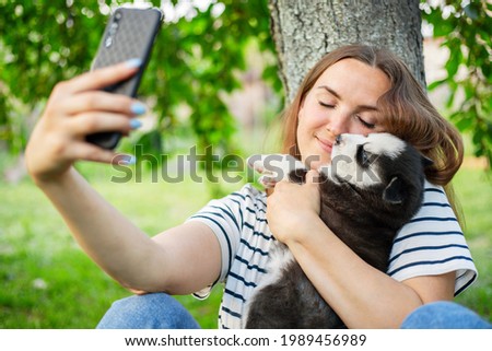 Young beautiful woman in a t-shirt and jeans with a husky puppy takes a photo or selfie on the phone while sitting in a park near a tree.