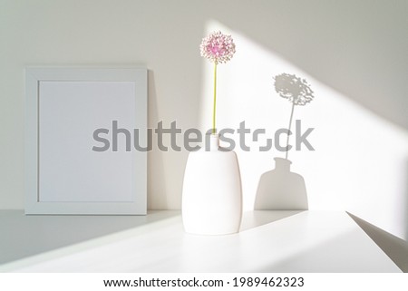 Empty white picture frame mockup. Modern and elegant vase with allium ampeloprasum flower in sunlight with long shadows. Minimal and white interior design. Minimalist and simple decoration