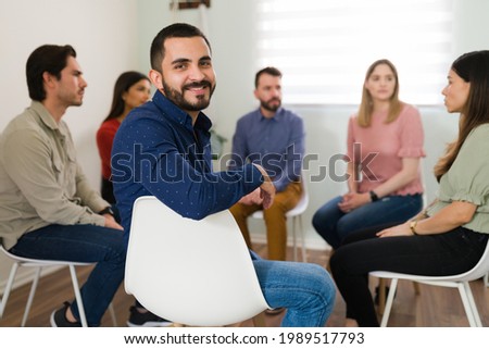 Portrait of a happy young man smiling and making eye contact while sitting during a meeting of support group. New member of an anger management group Royalty-Free Stock Photo #1989517793