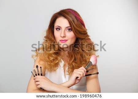 Young beautiful woman with bright pink make up and curly hair holding makeup brushes in her hands and posing on grey background at studio. Visagiste, makeup artist.