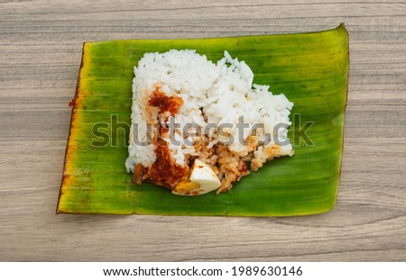 Flatlay picture of "Nasi Lemak Seringgit' on wooden table. Coconut rice serve with sauce, nuts and anchovies wrap in banana leaf that cost one 1 Ringgit.
