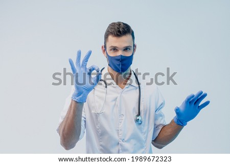 European male doctor in protective mask and latex gloves showing Ok gesture. Young man with stethoscope wearing white coat. Isolated on gray background with turquoise light. Studio shoot. Copy space.