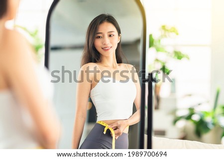  young woman takes a paper measuring ruler and measures her waist size before mirror. She is very satisfied with the results of the weight loss and the current figure. Royalty-Free Stock Photo #1989700694