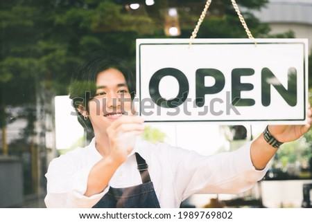 Young asian man Flip the open label to welcome customer and open the coffee shop in morning. Male asian Barista open the coffee cafe at doorway with reflection in mirror.