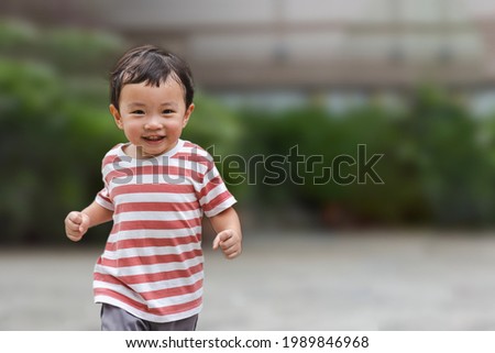 Portrait of an asian boy (toddler) running toward and smiling with happy and fun face while playing outdoor. A Child wear striped shirt in red and white color. Head and hair is wet by sweat. Royalty-Free Stock Photo #1989846968