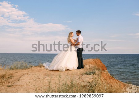 The bride and groom hug on the edge of the mountain against the background of the sea. An elegant bride in a white wedding dress looks lovingly at her husband. Happy newlyweds hugging