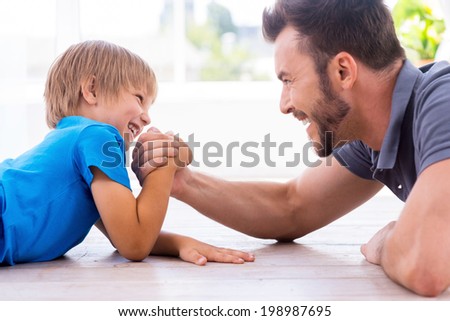 Little champion. Side view of happy father and son competing in arm wrestling while both lying on the hardwood floor 