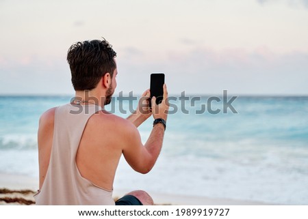 unrecognizable caucasian man with a cell phone in his hands taking pictures of the sunset at the sea of cancun, concept of technology.
