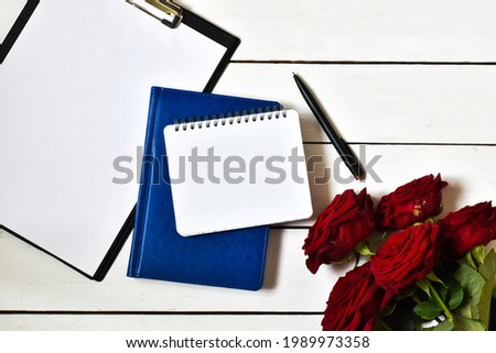 Bouquet of red roses with blank notepad, clipboard on a white wooden background. Greeting card. Flat lay, copy space. 