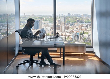 Indian serious investment banker financial analyst sitting at desk with papers working with big data, stock exchange trading operations using laptop near panoramic window in modern corporate office. Royalty-Free Stock Photo #1990075823