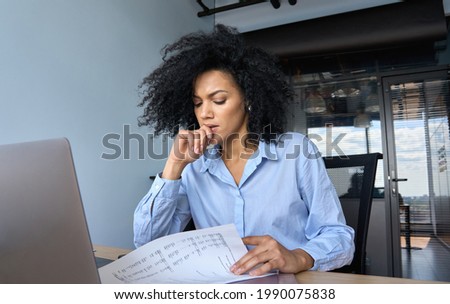 Concerned African American businesswoman ceo analyst sitting at desk reading papers financial reports with laptop in contemporary corporation office. Financial data analyst working with big data. Royalty-Free Stock Photo #1990075838