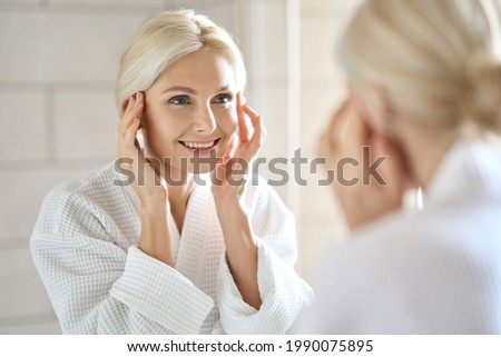 Gorgeous mid age adult 50 years old blonde woman standing in bathroom after shower touching face, looking at reflection in mirror smiling doing morning beauty routine. Older dry skin care concept. Royalty-Free Stock Photo #1990075895