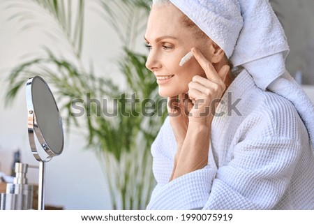 Gorgeous senior older adult 50 years old blonde woman wearing bathrobe and turban towel in bathroom applying moisturizing tightening face skin treatment, looking at mirror. Morning beauty routine. Royalty-Free Stock Photo #1990075919