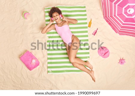 Top view of smiling relaxed curly haired woman with perfct figure lies on striped towel at beach listens music via headphones takes sunbath enjoys sun rests near sea dressed in bathing suit. Royalty-Free Stock Photo #1990099697