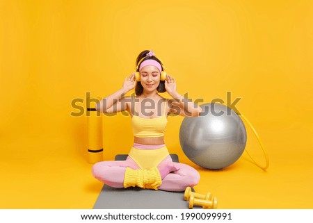 Fitness sport training concept. Sporty fit Asian woman sits crossed legs on mat enjoys listening music via headphones uses dumbbells fitball hula hoop has regular workout at home takes break