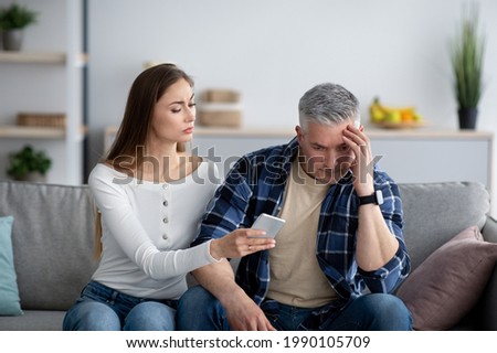 Mature woman confronting her husband about correspondence with his lover on smartphone. Middle-aged lady fnding out about her partner's love affair, accusing her spouse of marriage infidelity Royalty-Free Stock Photo #1990105709