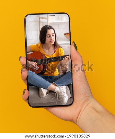 Female hand holding cellphone with online music lesson on screen, learning how to play guitar on web, orange background. Young guitarist teaching educational course on internet