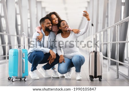Selfie At Airport. Happy Young Black Family Of Three People Taking Self Picture With Mobile Phone In Airport Terminal, Posing At Camera, Traveling Together. Daughter Embracing Parents From Behind