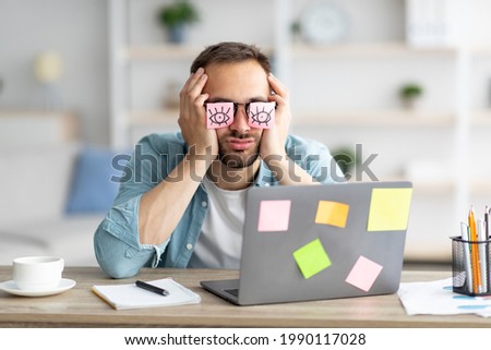 Tired young man having stickers with open eyes drawing on his glasses, sleeping in front of laptop at home office. Exhausted millennial man overworking, napping, failing to meet deadline