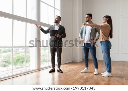 Professional Real Estate Agent Showing Spacious New Apartment To Young Couple, Looking And Pointing At Beautiful View From Big Panoramic Picture Windows. Happy Man And Woman Ready to Become Homeowners