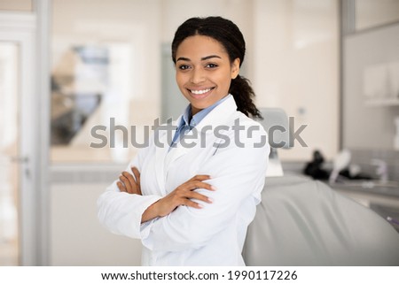 Portrait Of Beautiful Professional Black Female Stomatologist Posing In Clinic Interior, Happy African American Dentist Lady Standing With Folded Arms And Smiling At Camera, Ready For Check Up Royalty-Free Stock Photo #1990117226