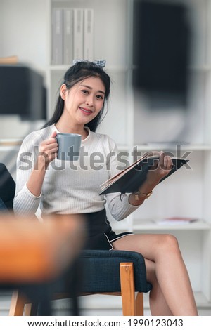Portrait of young beautiful asian woman holding cup and book while relaxing in living room, smiling and looking at camera.