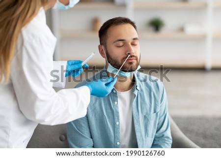 Doctor making covid-19 PCR test for male patient at home. Millennial guy undergoing coronavirus diagnostic procedure indoors. Viral disease prevention and treatment concept Royalty-Free Stock Photo #1990126760
