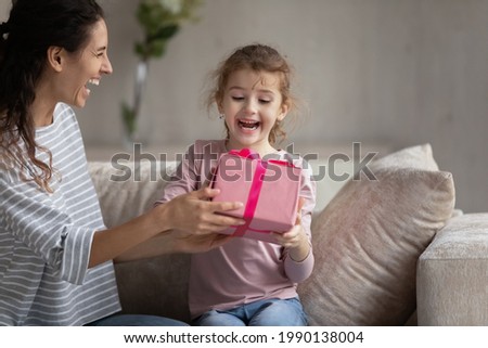 Smiling young Latin mom give wrapped gift box to excited small biracial daughter congratulate with birthday. Happy loving Hispanic mother make surprise present to overjoyed ethnic girl child at home.