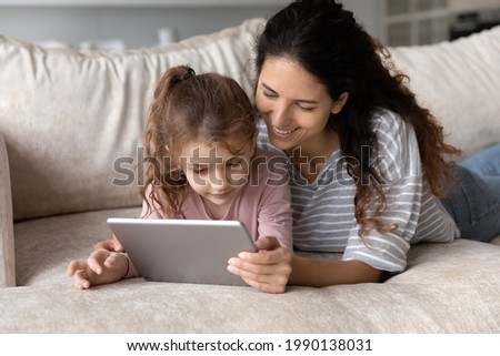 Happy loving young Latino mother and daughter lying relaxing on sofa at use modern tablet gadget together. Smiling Hispanic mom and small girl child look at pad screen browse internet on device.