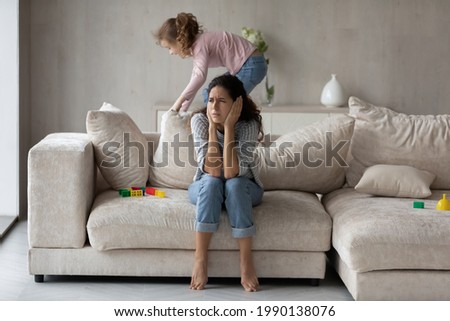 Exhausted Hispanic young mother bothered by loud noise biracial teen daughter playing at home. Tired Latin mom annoyed by ill-behaved naughty kid, suffer from headache. Parenthood concept. Royalty-Free Stock Photo #1990138076